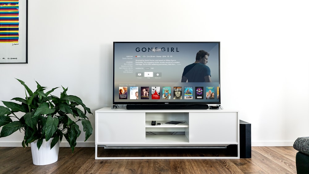 Smart TV ligada sobre um rack branco em uma sala de estar com planta ao lado
